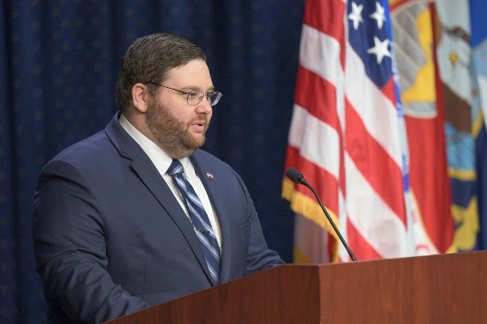 Person standing behind podium in front of flags wearing a suit with glasses.
