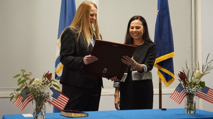 Ms Jennifer Desautel and Ms Ida Pham holding a frame together in front of two flags with a table in front of them