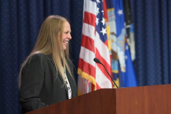 Person smiling wearing a black jacket standing behind a podium with long hair in front of flags.