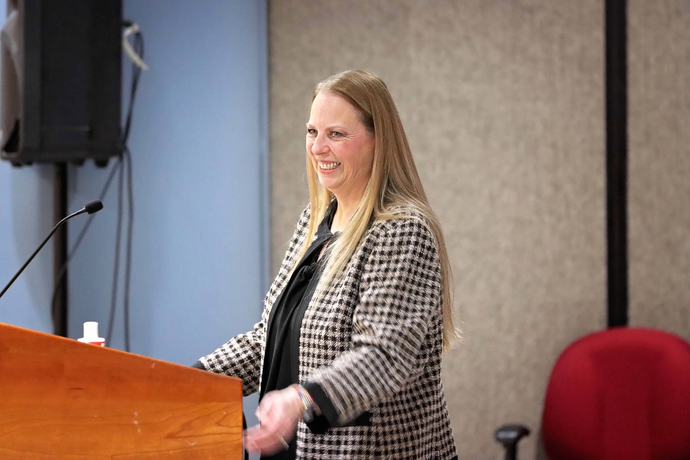 A woman standing at a podium smiling with a black and white jacket on in front of sound panels.