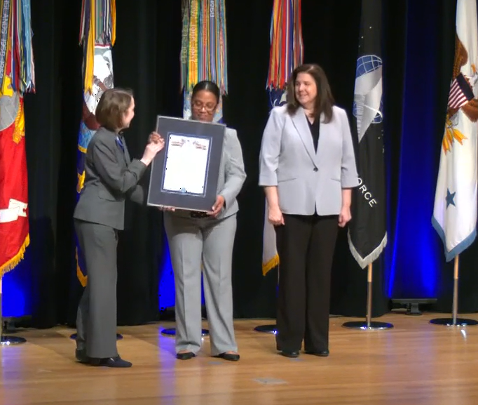 Three professionally dressed people stand on stage in front of colorful flags with an award. Three professionally dressed people stand on stage in front of colorful flags with an award.